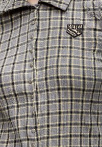 Gros plan d’une chemise à carreaux beige et noire avec des boutons-pression argentés et un patch de la marque Diesel sur la poitrine.