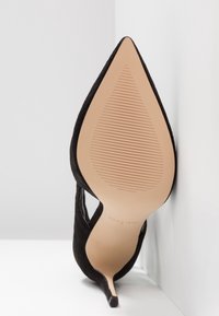Chaussure à talon haut en daim noir à bout pointu, inclinée vers le haut, montrant une semelle beige lisse avec des lignes de grip fines et un embout de talon mince.