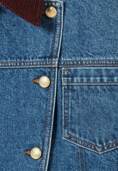 Sezione di giacca di jeans con colletto in velluto a coste di un rosso scuro, bottoni dorati, tessuto in denim azzurro chiaro, cuciture visibili e taschino sul petto.