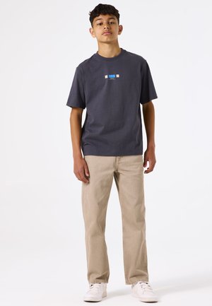 Camiseta de algodón gris con un pequeño diseño gráfico azul y beige; combinada con pantalones rectos beige claros y zapatillas blancas.