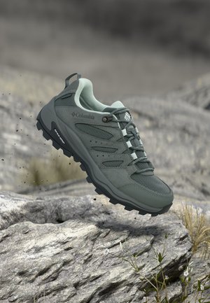 Chaussure de randonnée grise Columbia avec une semelle robuste suspendue au-dessus d'un terrain rocheux dans un cadre extérieur avec de l'herbe sèche et des fleurs.