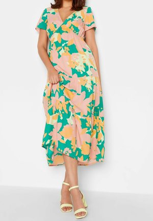 Femme portant une robe midi fleurie aux couleurs pastel rose, verte et jaune, manches courtes, et des talons ouverts jaunes à bride cheville, debout sur fond blanc.