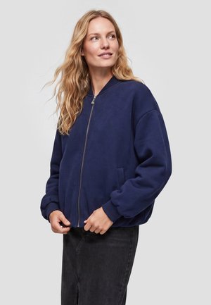 Donna bionda con giacca blu navy con zip e gonna nera, guarda di lato con le mani vicino alla zip della giacca.