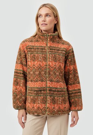 Kvinde iført en højhalset fleecejakke med lynlås og orange, brune og grønne geometriske mønstre, stående mod en ensfarvet baggrund.