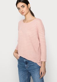 Suéter malha cor-de-rosa claro com riscas horizontais, decote largo e ombros caídos, combinado com jeans azuis de cintura alta.