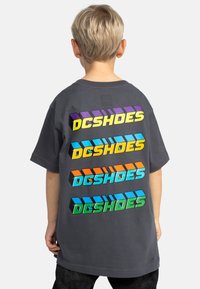 Šedé tričko s vícebarevným nápisem "DCSHOES" tištěným vertikálně na zadní straně v purpurové, žluté, modré, oranžové a zelené. Krátké rukávy.