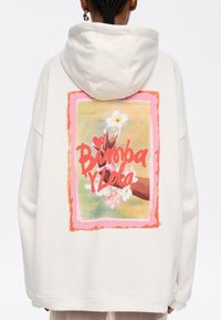 Sudadera de cuello redondo en algodón color crema, presenta un gráfico colorido en la parte posterior con flores y texto manuscrito. Diseño con capucha, ajuste casual.