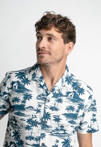 Chemise à manches courtes à boutons blanche avec motif tropical bleu, comportant des palmiers, des bateaux et des silhouettes de bâtiments. Texture lisse.