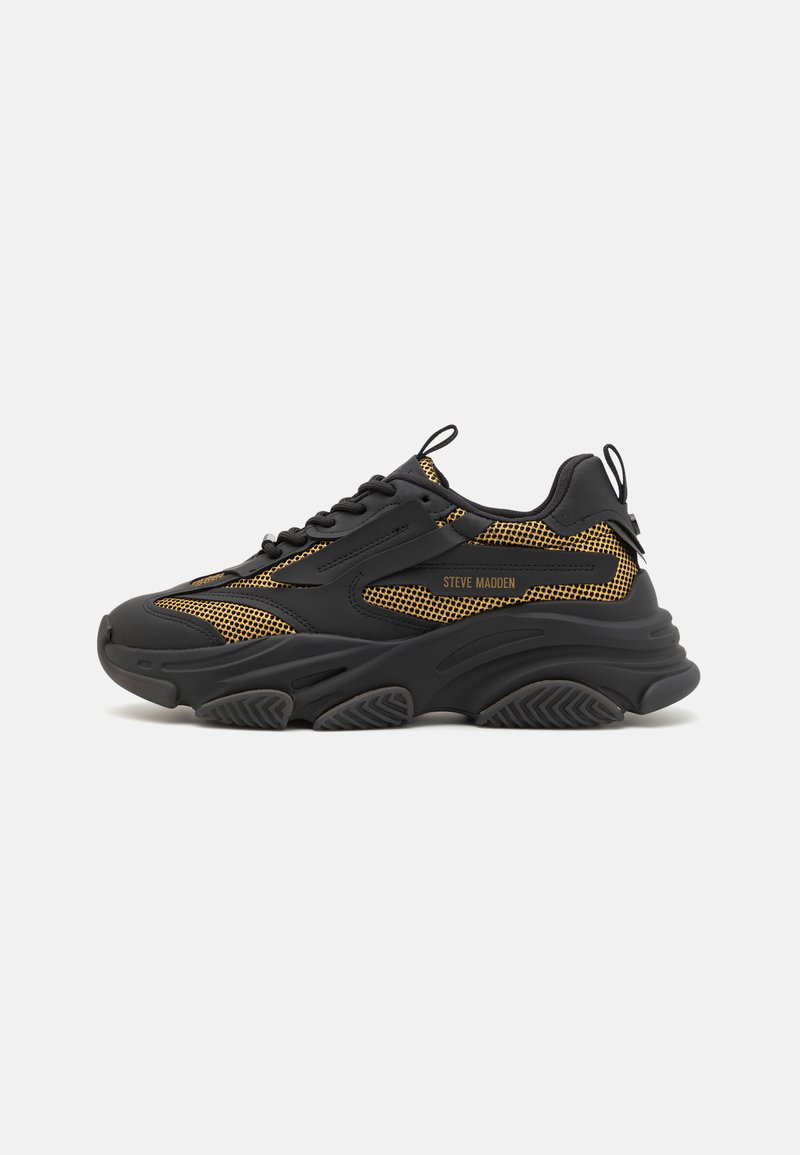 Zapatillas deportivas negras y doradas con parte superior de malla, suela de goma gruesa, cordones y tira de agarre. Presenta acentos texturizados y detalles del logo.