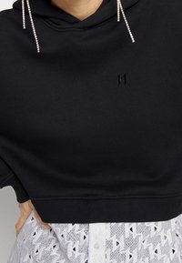 Sudadera negra corta con capucha y logo bordado; presenta acentos de strass que cuelgan de la capucha. Combinada con una parte superior blanca con estampado.
