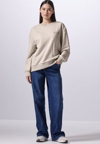 Beige sweatshirt met een ronde hals en grote cuffs, gecombineerd met wijde blauwe jeans. Tekstdetail in bruin op de borst.