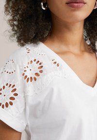 Witte V-hals top met geborduurde floral uitsneden op de mouwen. Zachte katoenen stof met een gladde textuur en een relaxte pasvorm.