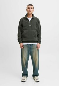 Young man standing wearing a dark green quarter-zip fleece, faded blue jeans, white sneakers, and a white t-shirt underneath.