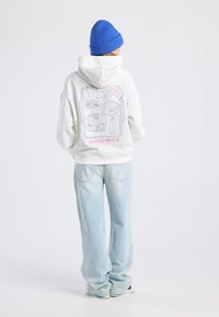 Person wearing white hoodie with breakfast tray illustration and "ROOM SERVICE" text on back, blue beanie, light blue jeans, and sneakers.