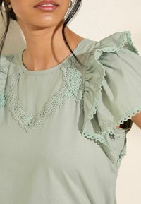 Top verde chiaro con maniche arruffate e bordo in pizzo attorno al collo. Tessuto morbido con delicati accenti in pizzo. Texture e dettagli di design sottili.