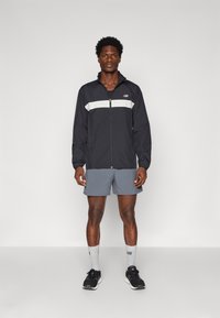 Black zip-up windbreaker with gray horizontal stripe, paired with gray shorts. Worn with black sneakers and gray socks. Simple design.