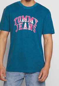 Camiseta de algodón color teal con cuello redondo, que presenta el logotipo en blanco y rosa "TOMMY JEANS" en letras gruesas en la parte frontal. Mangas cortas.