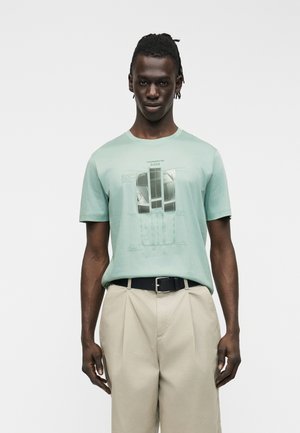 Mężczyzna ubrany w miętowo-zieloną koszulkę z nadrukiem samochodu, beżowe spodnie z plisami i czarny pasek, stojący na tle jednolitej, białej ściany.