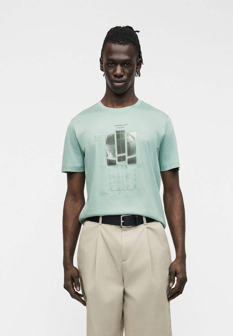 Άνδρας που φοράει ένα T-shirt σε απαλό πράσινο με σχέδιο αυτοκινήτου, μπεζ πλισέ παντελόνι και μαύρη ζώνη, στέκεται μπροστά σε απλό λευκό φόντο.