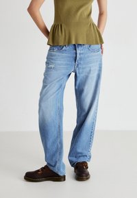 Jeans de pierna ancha en azul claro con una textura desgastada y ligero desgarro. Combinados con una blusa peplum verde y zuecos marrones.