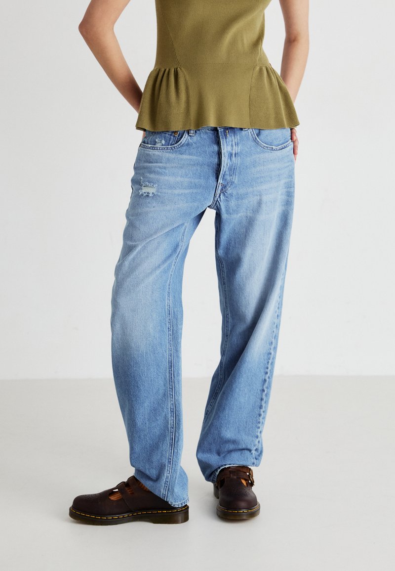 Jeans de pierna ancha en azul claro con una textura desgastada y ligero desgarro. Combinados con una blusa peplum verde y zuecos marrones.