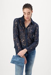 Marineblauwe kanten blouse met kraag, doorzichtige structuur en lange mouwen. Houdt een blauwe suède handtas vast, in combinatie met lichte spijkerbroeken.