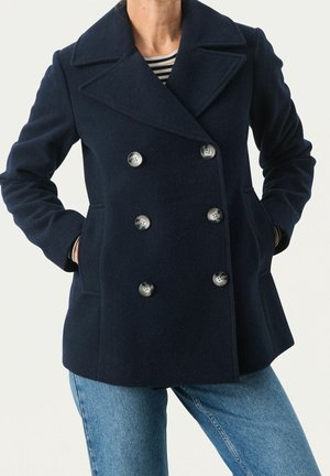 Manteau peacoat bleu marine croisé avec six boutons, porté sur une chemise à rayures et un jean bleu, mains dans les poches du manteau.