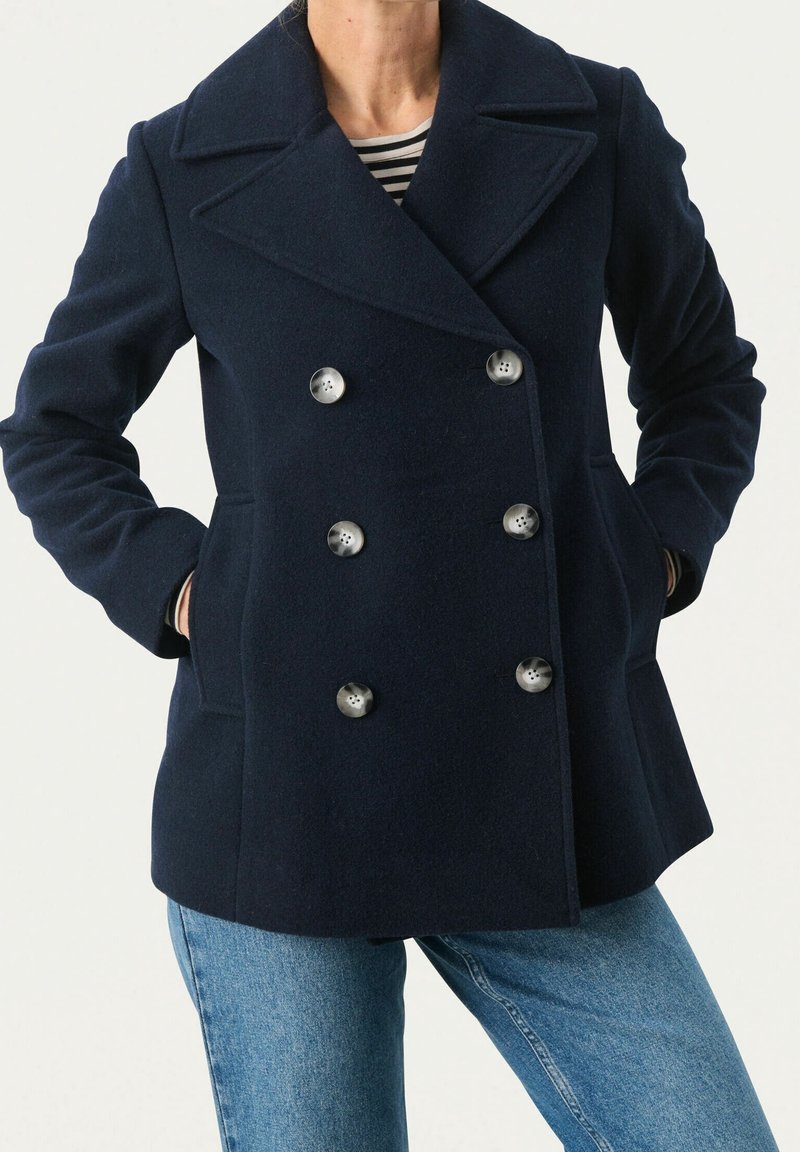 Manteau peacoat bleu marine croisé avec six boutons, porté sur une chemise à rayures et un jean bleu, mains dans les poches du manteau.
