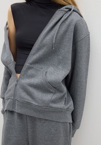 Grijze zip-up hoodie van geborsteld materiaal, met een capuchon, voorzakken en geribbelde mouwen. Gedeeltelijk open gedragen over een zwart aansluitend topje.