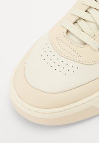 Beige bőr sportcipő sima felülettel, perforált orr résszel, lapos fűzőkkel és texturált gumitalppal.