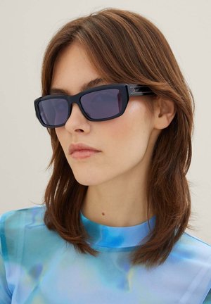 Occhiali da sole rettangolari neri con lenti scure. Montatura spessa con riflessi lucidi. Il modello indossa una maglietta blu a fantasia. Capelli lisci.