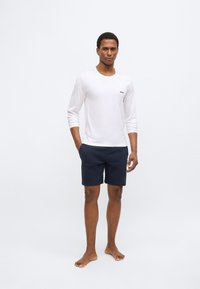 White long-sleeve shirt with a small logo, paired with navy shorts; smooth fabric; plain design; model standing barefoot.