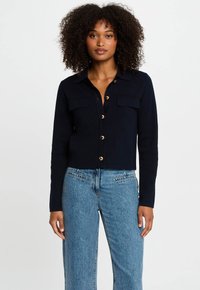 Marineblauwe cropped cardigan met knoopsluiting, twee voorzakken en goudkleurige knopen. Zachte stof met een gladde textuur. Gepaard met lichtblauwe spijkerbroek.