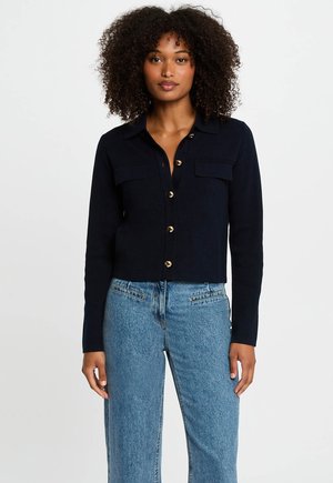 Cardigan corto navy con chiusura a bottoni, due tasche frontali e bottoni dorati. Tessuto morbido con una texture liscia. Abbinato a jeans azzurri chiari.