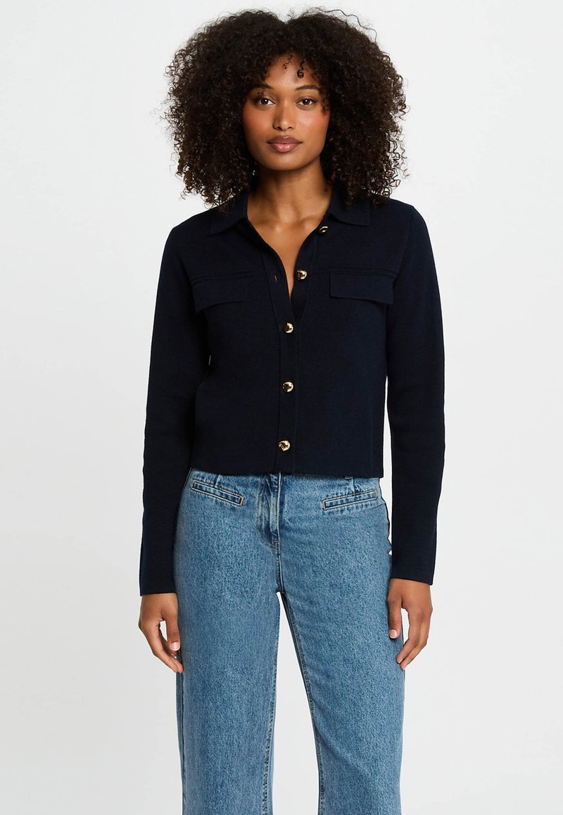 Marineblauwe cropped cardigan met knoopsluiting, twee voorzakken en goudkleurige knopen. Zachte stof met een gladde textuur. Gepaard met lichtblauwe spijkerbroek.