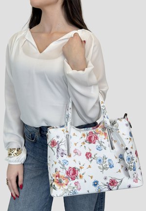 Kvinde iført hvid bluse og blå jeans holder en hvid lædertaske med farverigt blomsterprint og to håndtag.