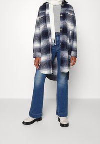 Námořnický a bílý kostičkovaný oversize kabát se dvěma předními kapsami, kombinovaný s modrými rozšířenými džíny a krémovými kotníkovými botami. Měkká textura a detaily s knoflíky.