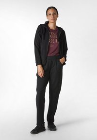 Felpa nera con zip, t-shirt bordeaux con testo dorato e pantaloni neri. L'outfit ha una vestibilità comoda e tasche laterali, abbinato a sneakers nere.