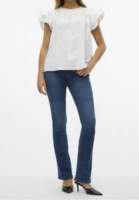 Donna che indossa una blusa bianca a maniche corte con volant, jeans blu a gamba dritta e scarpe nere con tacco alto su uno sfondo neutro.