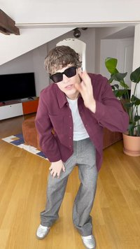 Maron rød skjorte med knapper, hvit T-skjorte, grå rutete bukser og sølvfargede joggesko. Sorte solbriller. Innendørs omgivelser med planter og en TV.