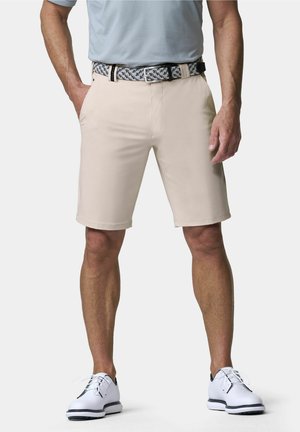 Mann trägt beige Shorts, gewebten schwarz-weißen Gürtel, hellblaues Hemd und weiße Golfschuhe, steht mit den Händen in den Taschen.