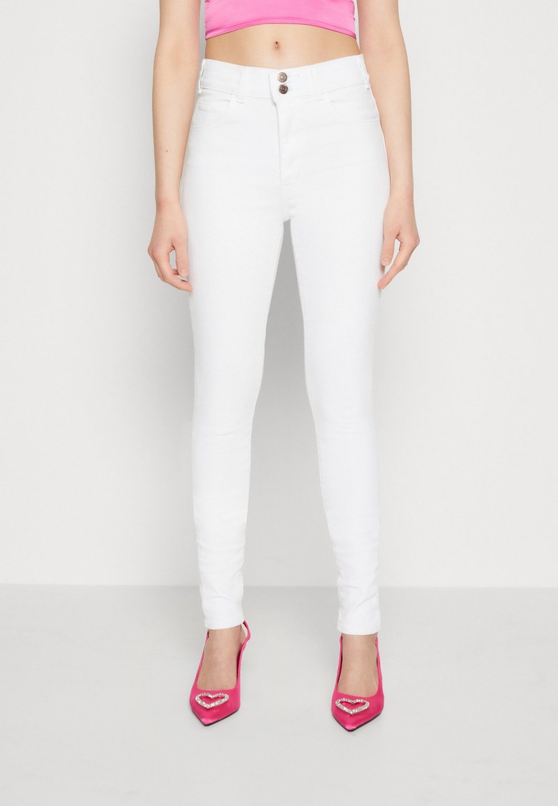 Pantalones vaqueros blancos ajustados con cintura alta y cierre de dos botones, combinados con zapatos de tacón altos de color rosa brillante que presentan acentos en forma de corazón.