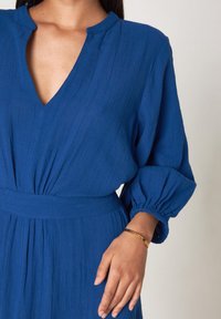 Robe bleue avec un décolleté en V profond, de longues manches bouffantes et un tissu texturé ; présente une taille cintrée et une silhouette décontractée.