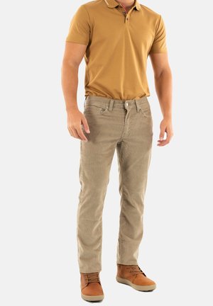 Pantalones de pana beige con un corte ajustado, cinco bolsillos y cierre frontal, combinados con una camiseta polo mostaza y botas marrones para un look casual.