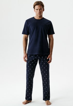 Mężczyzna stojący boso, ubrany w granatowy t-shirt z krótkim rękawem i ciemne wzorzyste spodnie od piżamy na jednolitym białym tle.