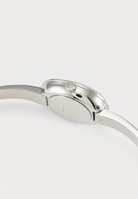 Montre argentée avec un boîtier circulaire en acier inoxydable, un bracelet lisse et une couronne avec un logo. Elle présente un design minimaliste avec une finition polie.