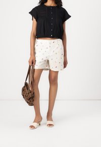 Blusa nera a maniche corte con chiusura a bottoni, abbinata a pantaloni corti bianchi con motivi floreali colorati, mule beige con punta aperta e borsa marrone.