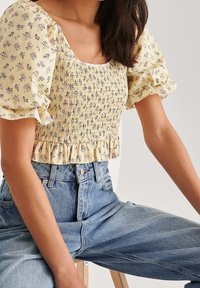 Top court floral jaune avec des manches bouffantes, présentant un design smocké et un ourlet volanté. Porté avec un jean en denim taille haute.