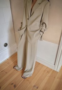 Bežna oversize jakna sako z žepom in gumbom, v kombinaciji s širokimi hlačami. Gladka tekstura, obuta v črne sandale na lesenem tleh.