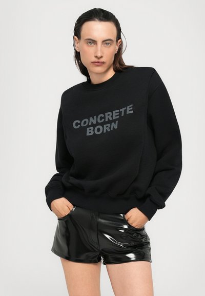 Felpa nera con la scritta "CONCRETE BORN" stampata in tonalità scura, abbinata a pantaloni corti neri lucidi. Il tessuto è liscio con una vestibilità rilassata.